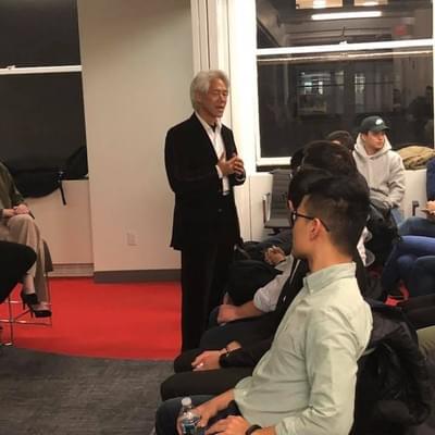 A speaker in a dark suit addressing a seated audience at an evening Harvard Alumni in Tech talk