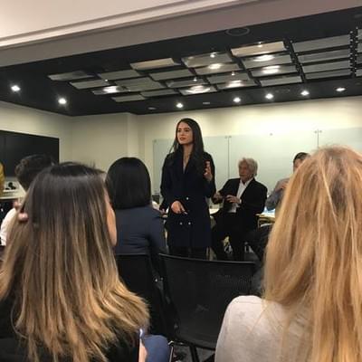 A speaker in a navy blazer standing to address a seated audience in a conference room