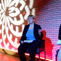 A panelist speaking on a red-lit stage in front of a patterned brick backdrop