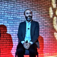 A bearded panelist seated alone on stage against a red and blue lit brick wall