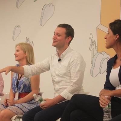 Three panelists seated in front of a cloud-patterned wall, one gesturing while speaking