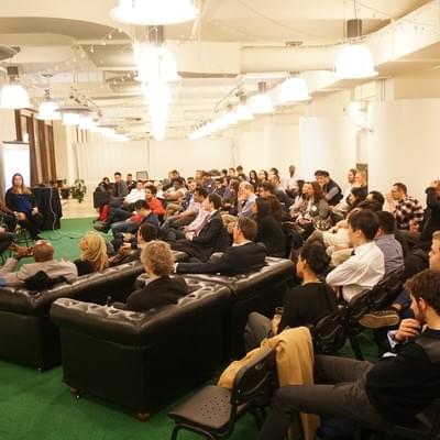 Attendees seated on sofas and chairs on green carpet, listening to a speaker