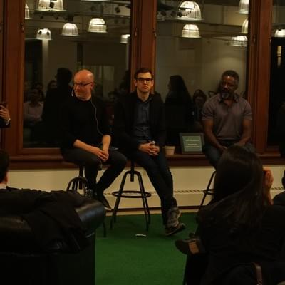 Three panelists on stools taking questions from an audience seated on green carpet