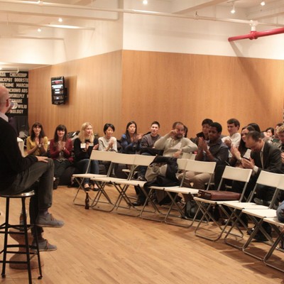 A speaker on a stool addressing attendees seated in rows of folding chairs
