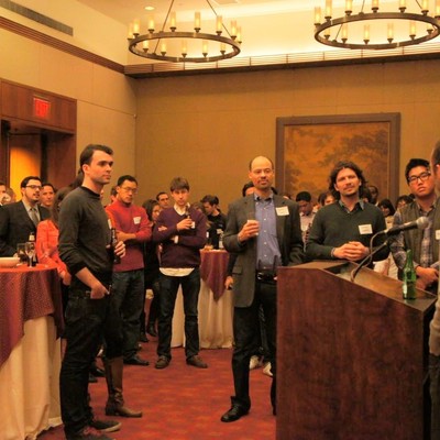 Attendees standing with drinks, listening to remarks given from a podium