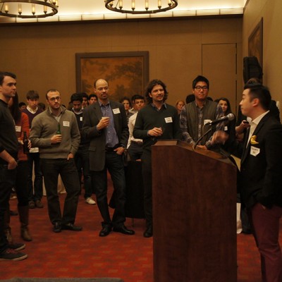 A speaker giving remarks at a wooden podium while attendees stand and listen