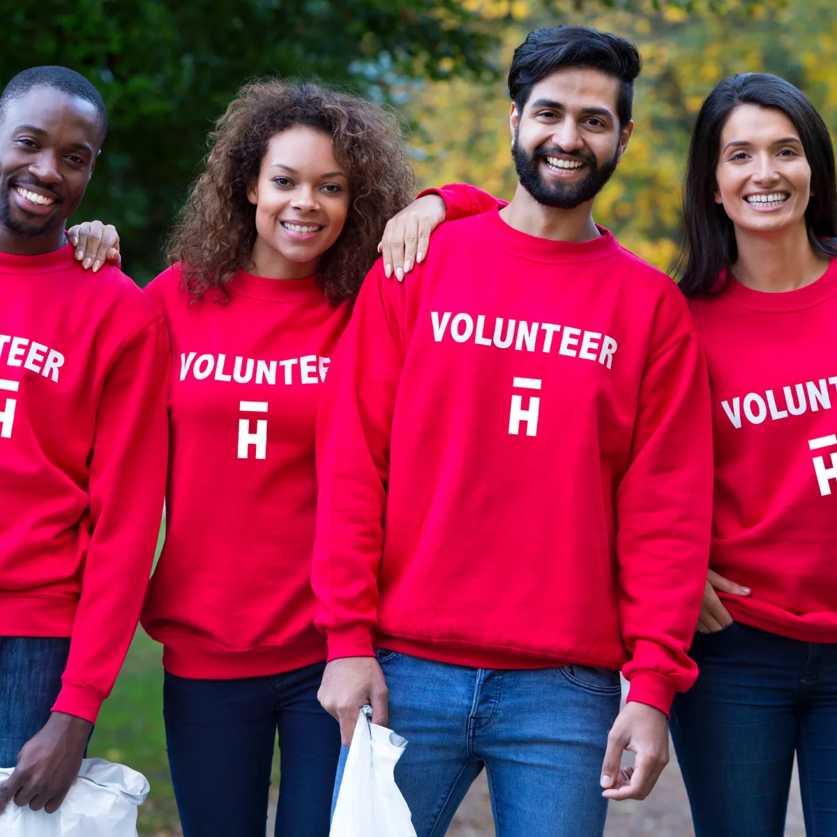 Four smiling volunteers in red VOLUNTEER sweatshirts standing arm in arm outdoors, two holding rubbish bags after a community clean-up